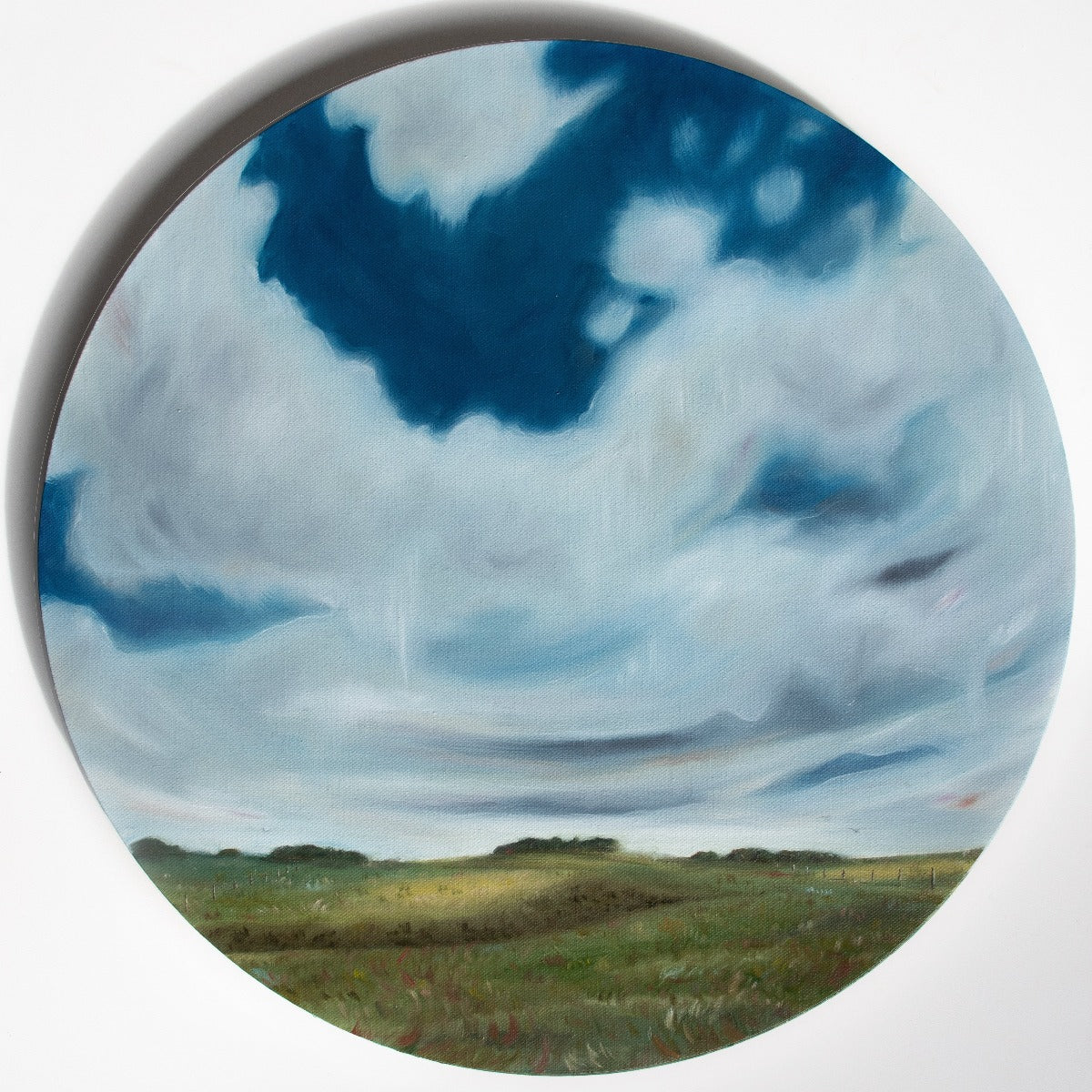 The road home is a round oil painting showing a dramatic soaring sky above a rolling farm landscape. The grasses are swept by the wind with a fenceline marching into the distance and trees on the horizon.